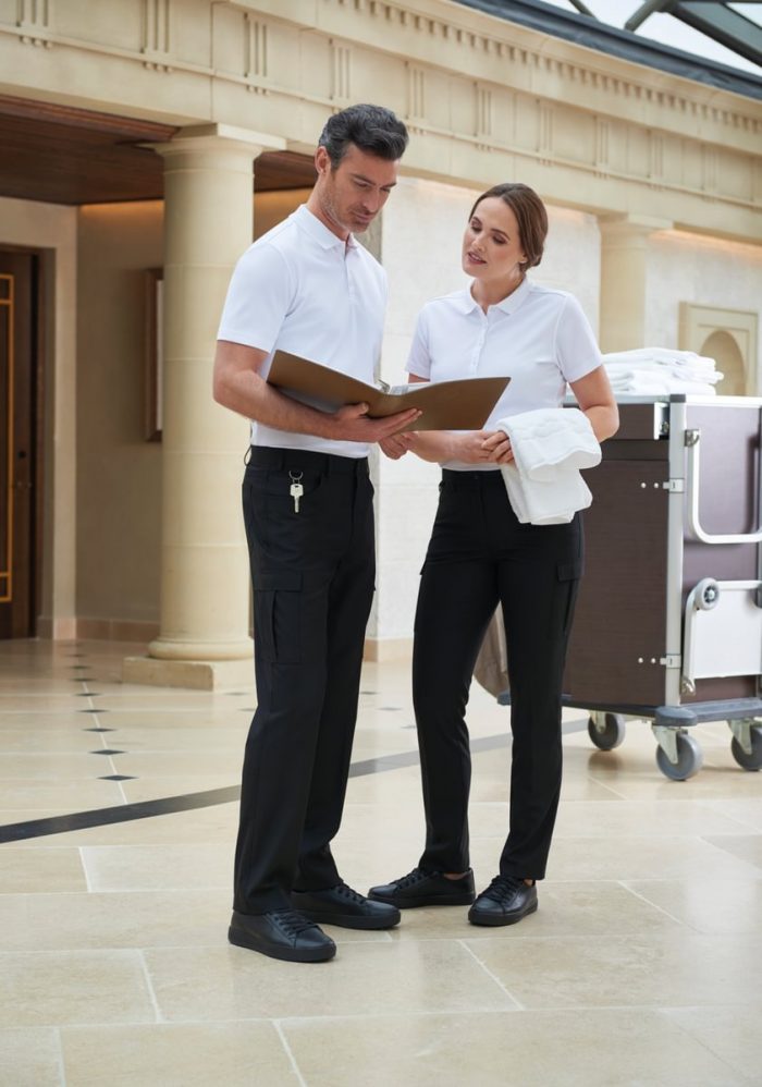 Two members of hotel staff, one male and one female, both in matching uniforms of a white polo and black trousers, looking over a ledger