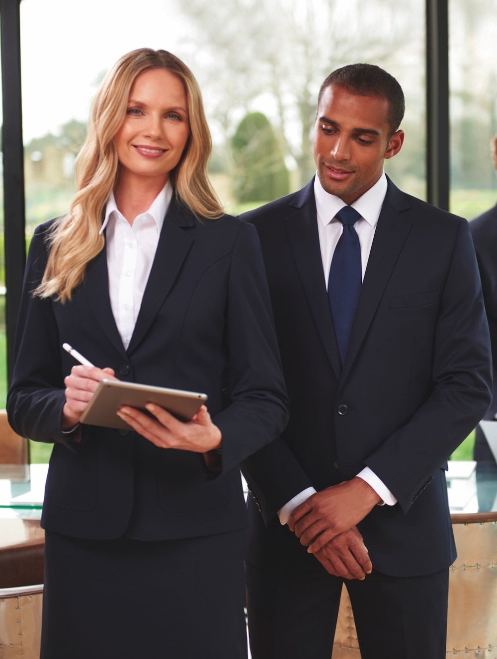 A man and a woman in black suits. The woman is working on a tablet while the man watches