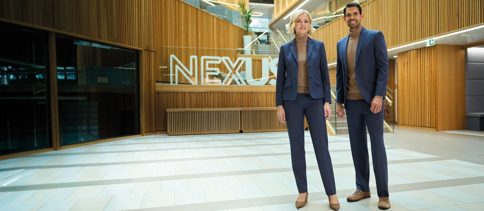 A man and a woman in blue suits and brown roll-neck jumpers standing in an office lobby