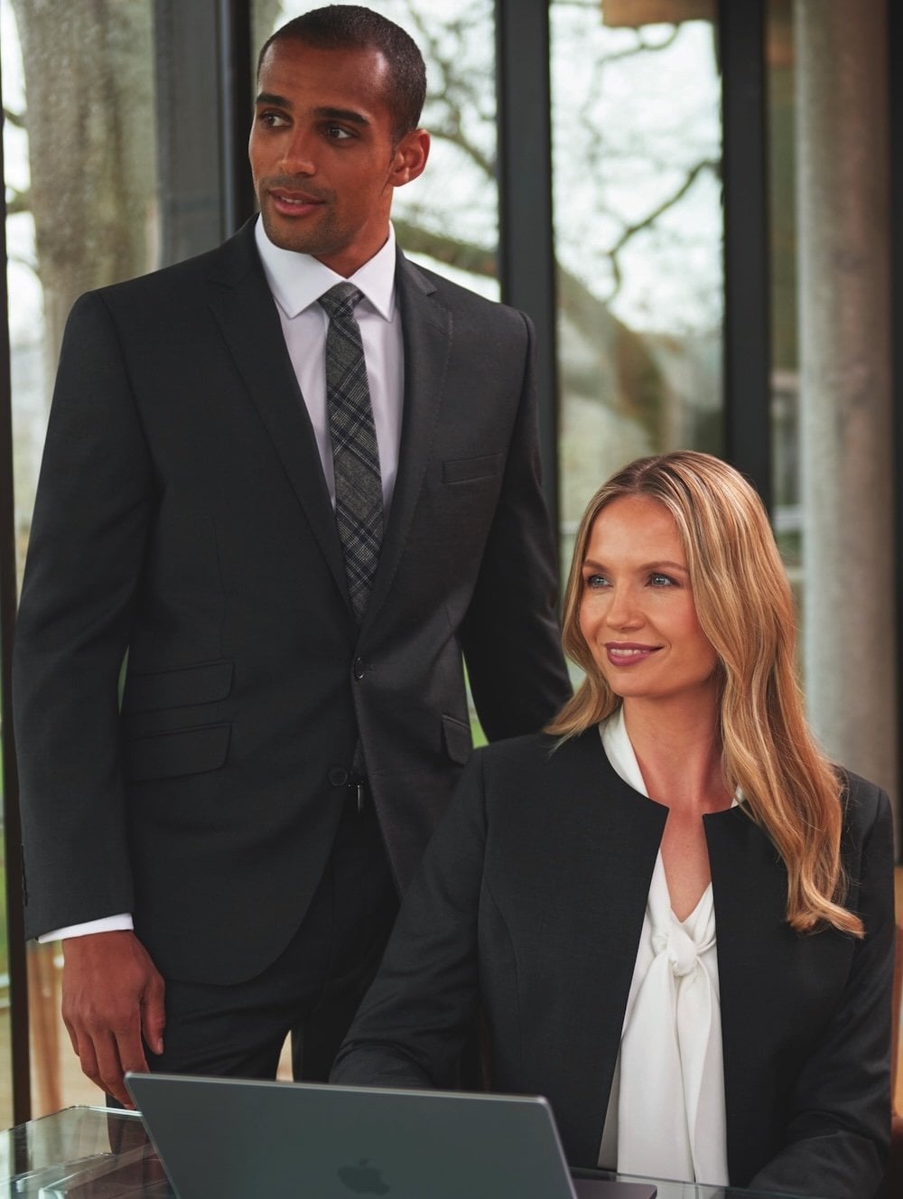 A man standing next to a woman on a laptop, both wearing black suits